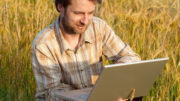 Feliz sonriente agricultor moderno de cuarenta años revisando su campo de trigo y trabajando en el ordenador portátil; Shutterstock ID 145449976; PO: AAF general